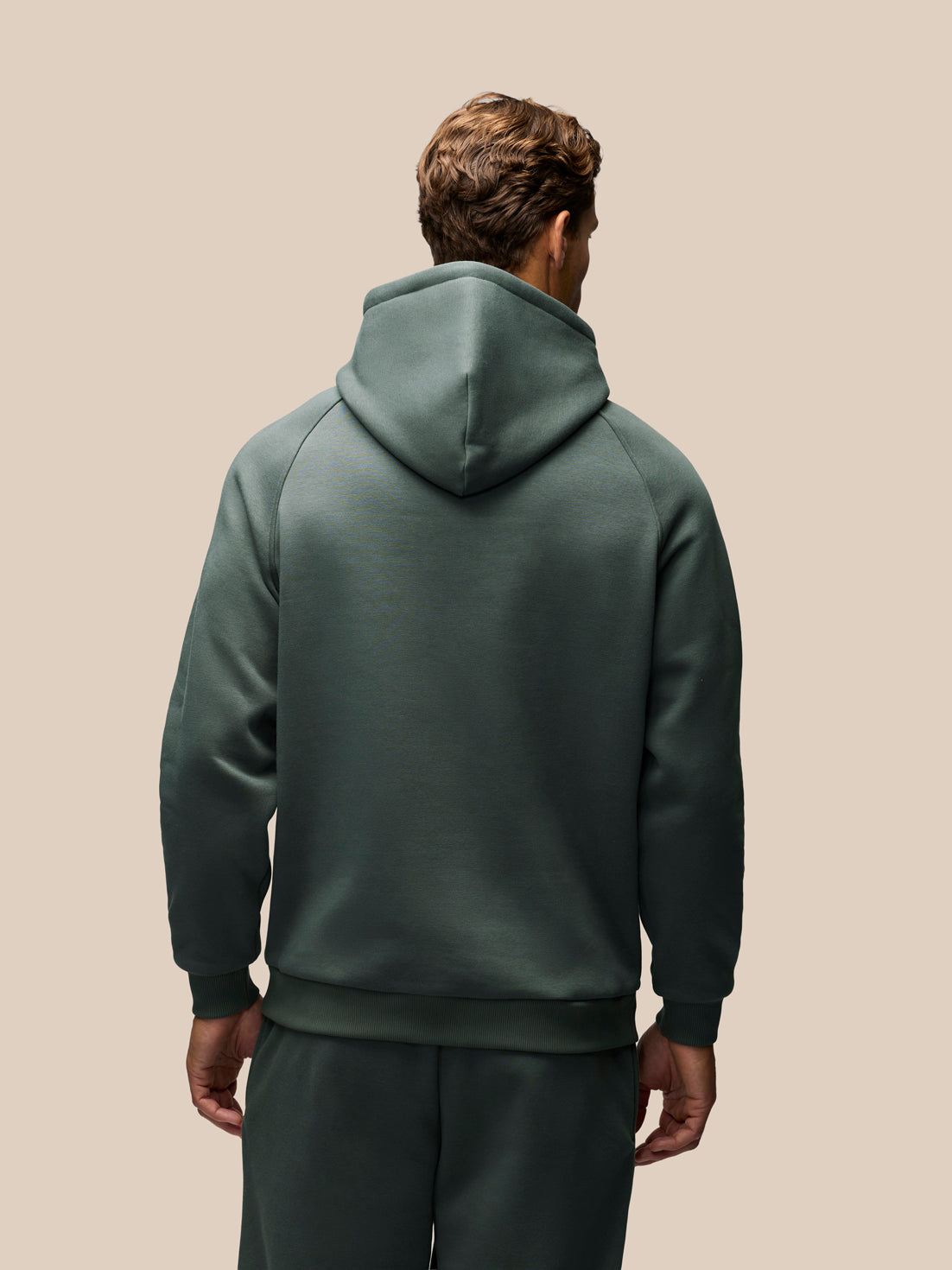 A man viewed from behind wearing a dark green hooded sweatshirt and matching sweatpants against a solid beige background.