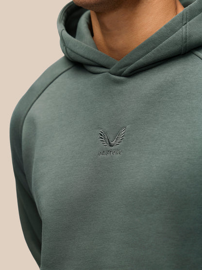 Close-up of a person wearing a muted sage green pullover hoodie with a tonal embroidered winged logo and the word 'CASTORE' on the chest, against a plain light beige background.