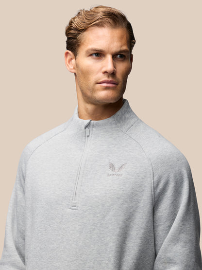 A man wearing a light gray heathered quarter-zip pullover sweatshirt with a small embroidered winged logo on the left chest, set against a solid beige background.