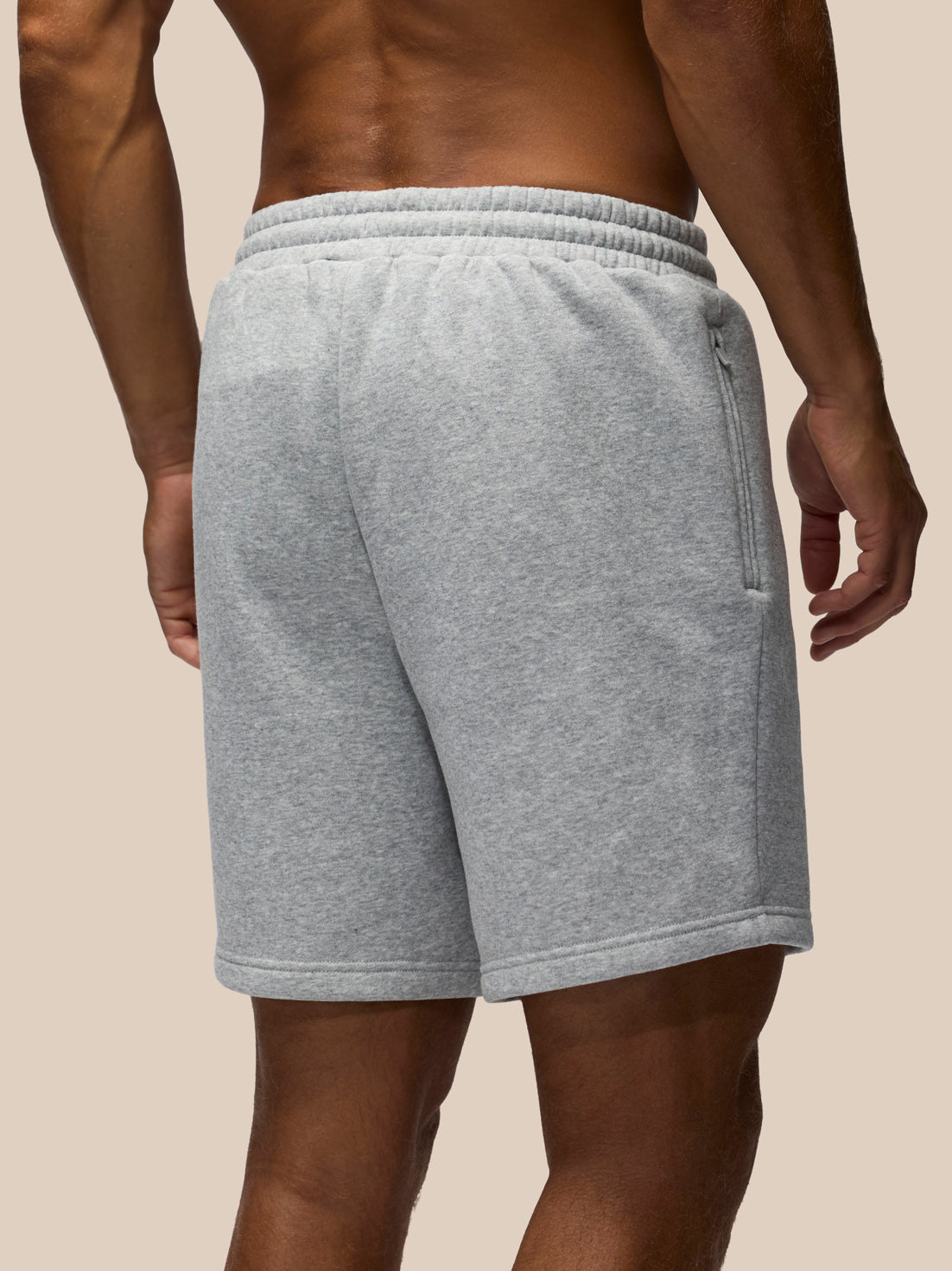 A close-up back view of a person wearing light heather gray sweat shorts against a solid beige background.