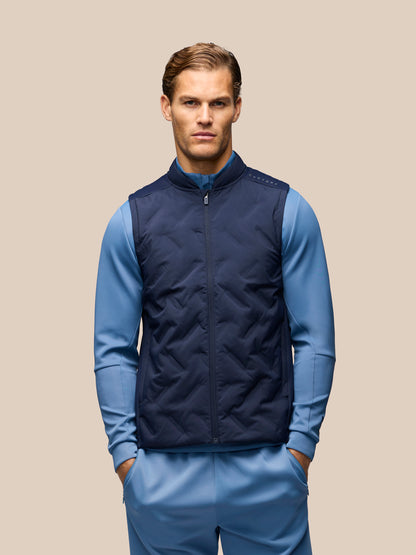 A male model wearing a navy blue quilted zip-up vest over a light blue long-sleeve top and matching blue pants, standing against a solid beige background.