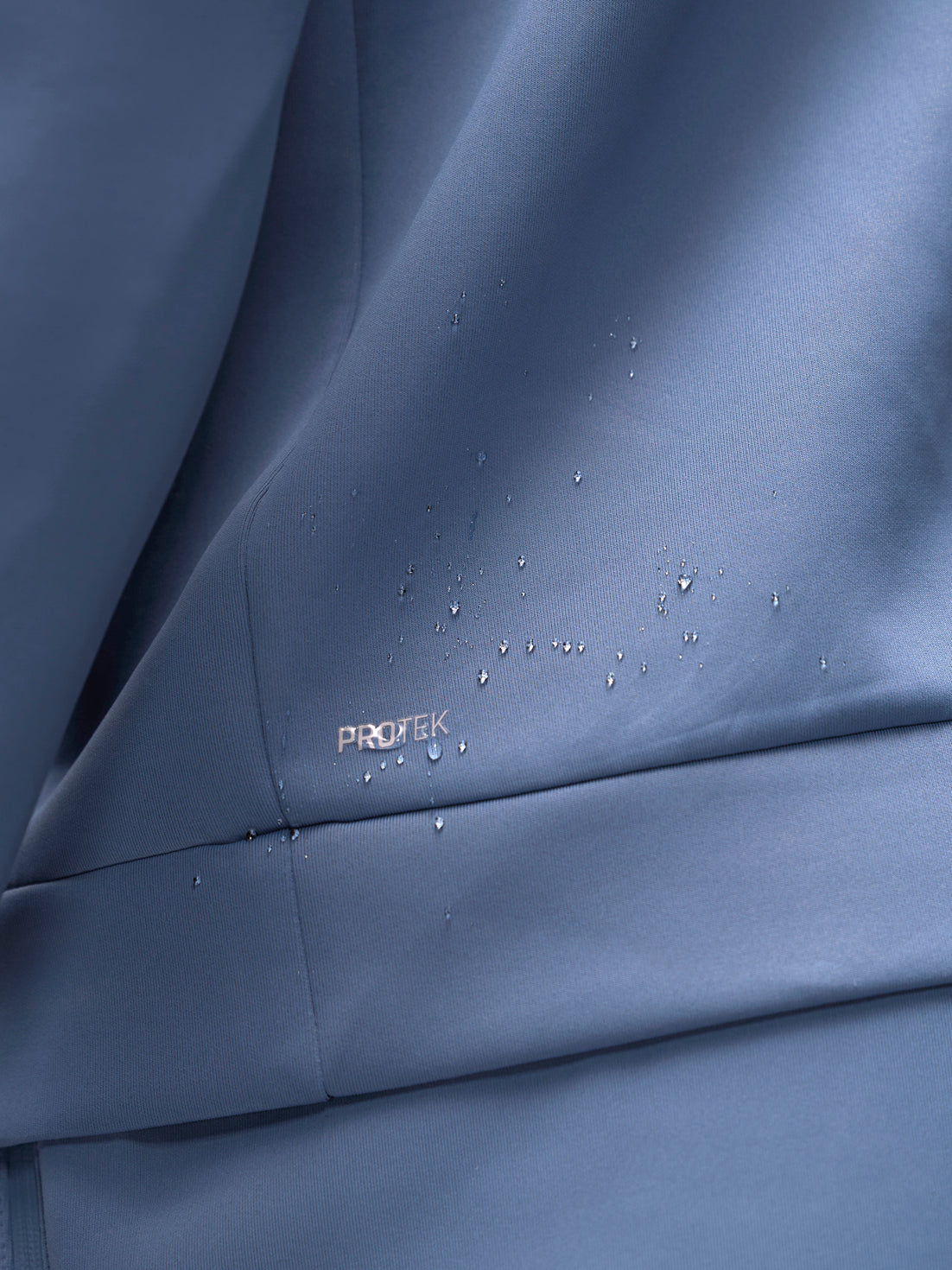Close-up of a blue-gray fabric surface with water droplets beading on it and a small visible PROTEK logo near the lower left area.