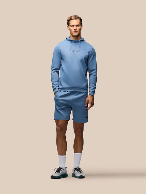 A male model stands against a solid beige background wearing a matching light blue athletic hoodie and shorts set, white crew socks, and gray running shoes.