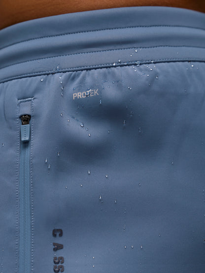 Close-up of a light blue athletic garment showing the PROTEK water-repellant finish with water droplets on the surface and a vertical zipper pocket.