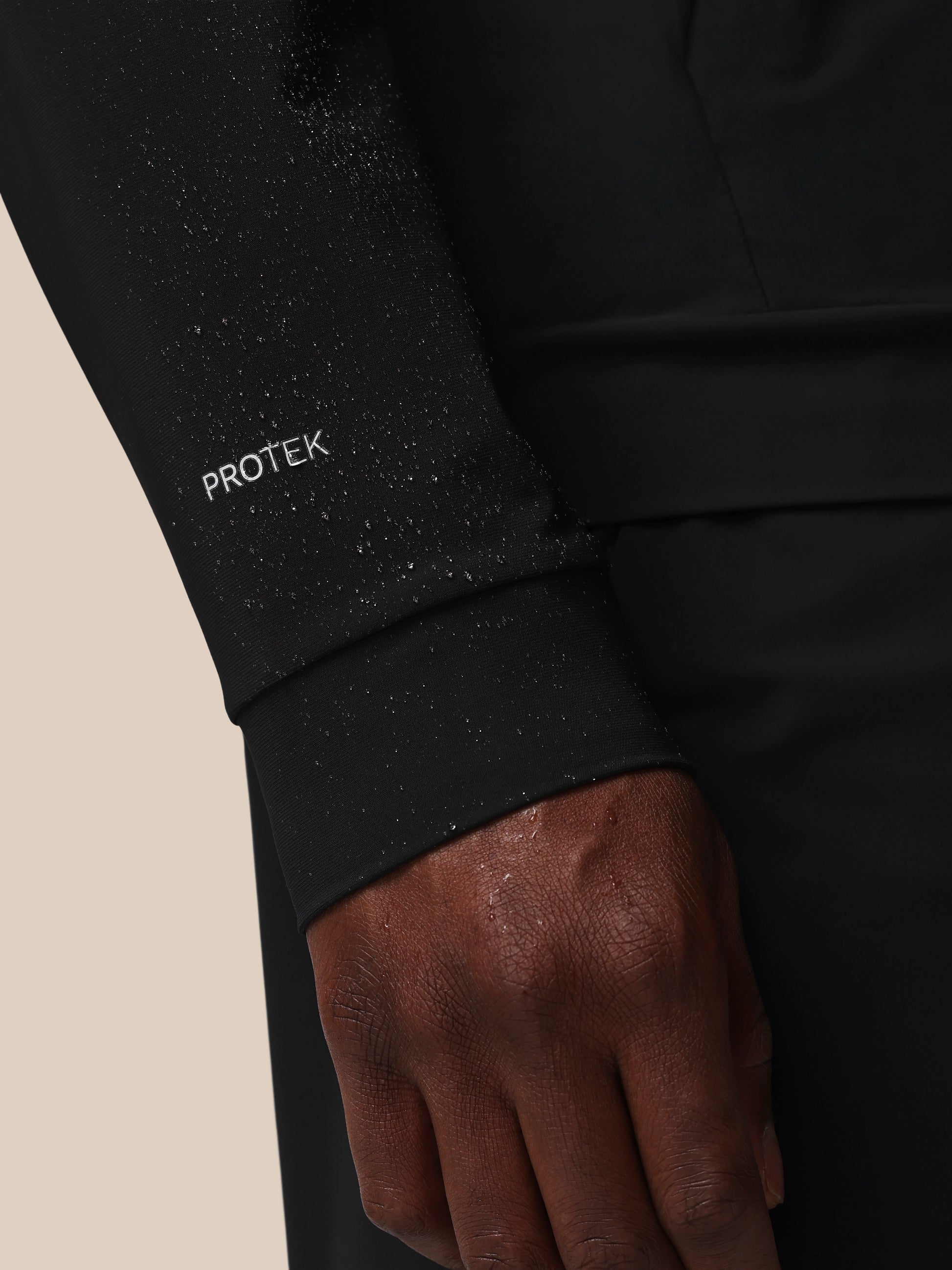 Close-up of black jacket sleeve with PROTEK label on the cuff and water droplets on the fabric.