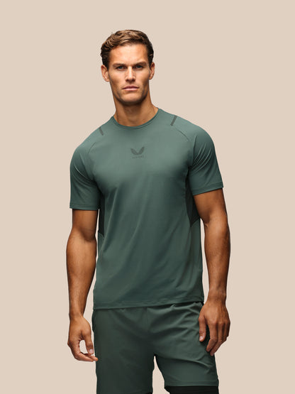 A man modeling a dark green athletic short-sleeve shirt and matching shorts against a solid beige background.