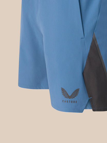 Close-up of a light blue athletic shorts leg with a dark gray mesh side panel and a dark gray Castore wing logo near the hem, set against a solid beige background.