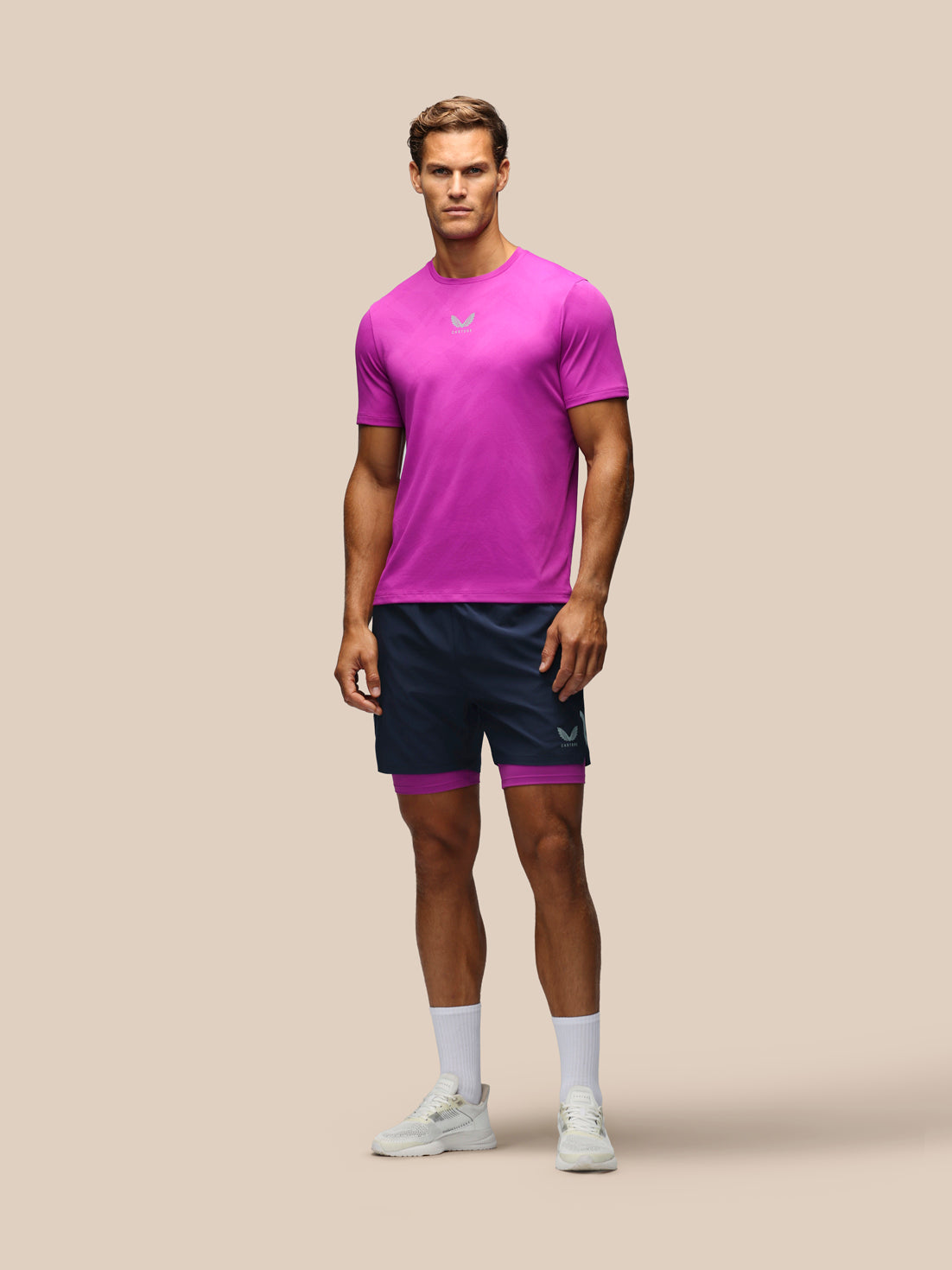 A male model stands against a solid beige studio background wearing a bright magenta athletic t-shirt, navy blue athletic shorts with a magenta inner lining, white socks, and white sneakers.