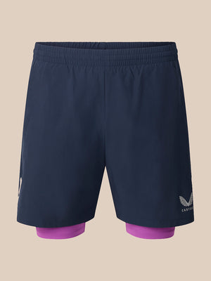 A pair of dark navy blue athletic shorts with a built-in purple inner layer visible at the hem, featuring a white wing logo and the brand name 'CASTORE' on the lower right leg, set against a solid beige background.