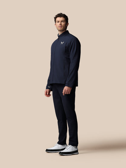 Male model wearing navy Castore Men’s Golf LVS Woven Jacket with zip front and chest logo against a beige backdrop.