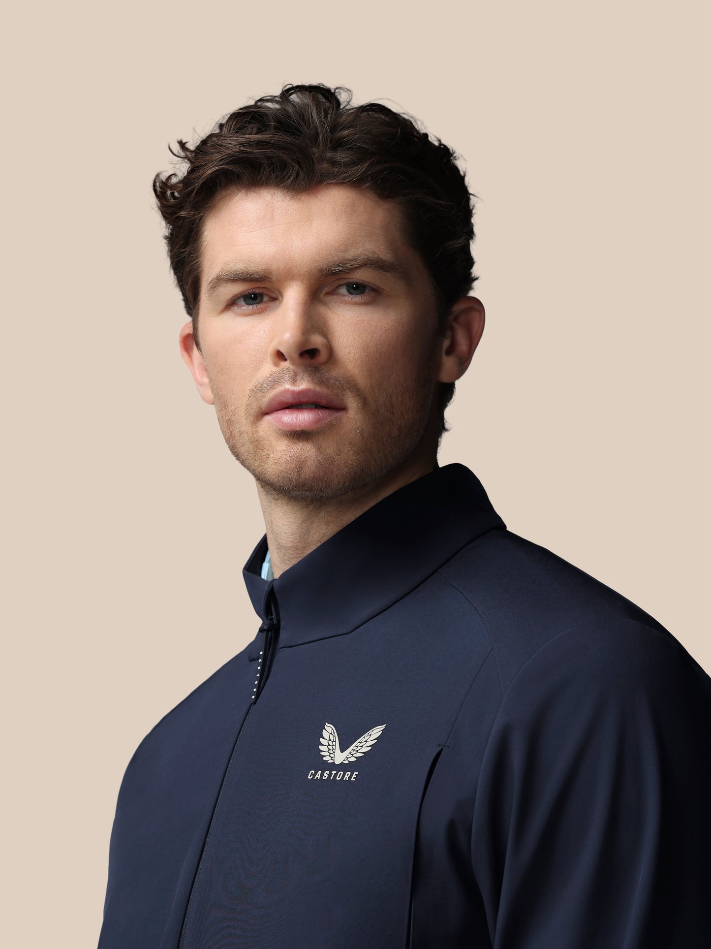 Man wearing navy Castore golf LVS woven jacket shown in a slightly angled front view with logo on chest against beige backdrop.