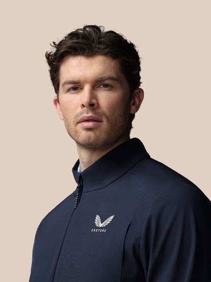 Man wearing navy Castore golf LVS woven jacket shown in a slightly angled front view with logo on chest against beige backdrop.