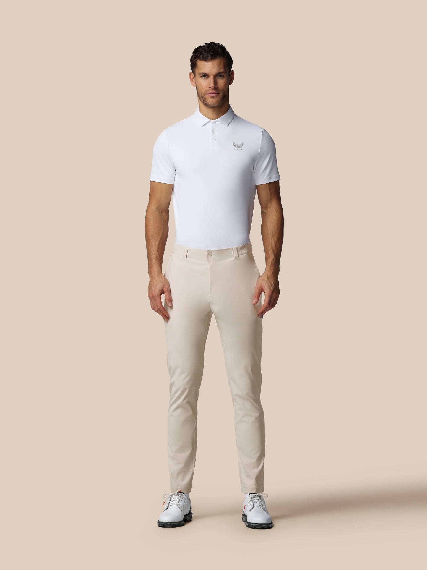 Male model wearing cream golf trousers and white polo against a beige background