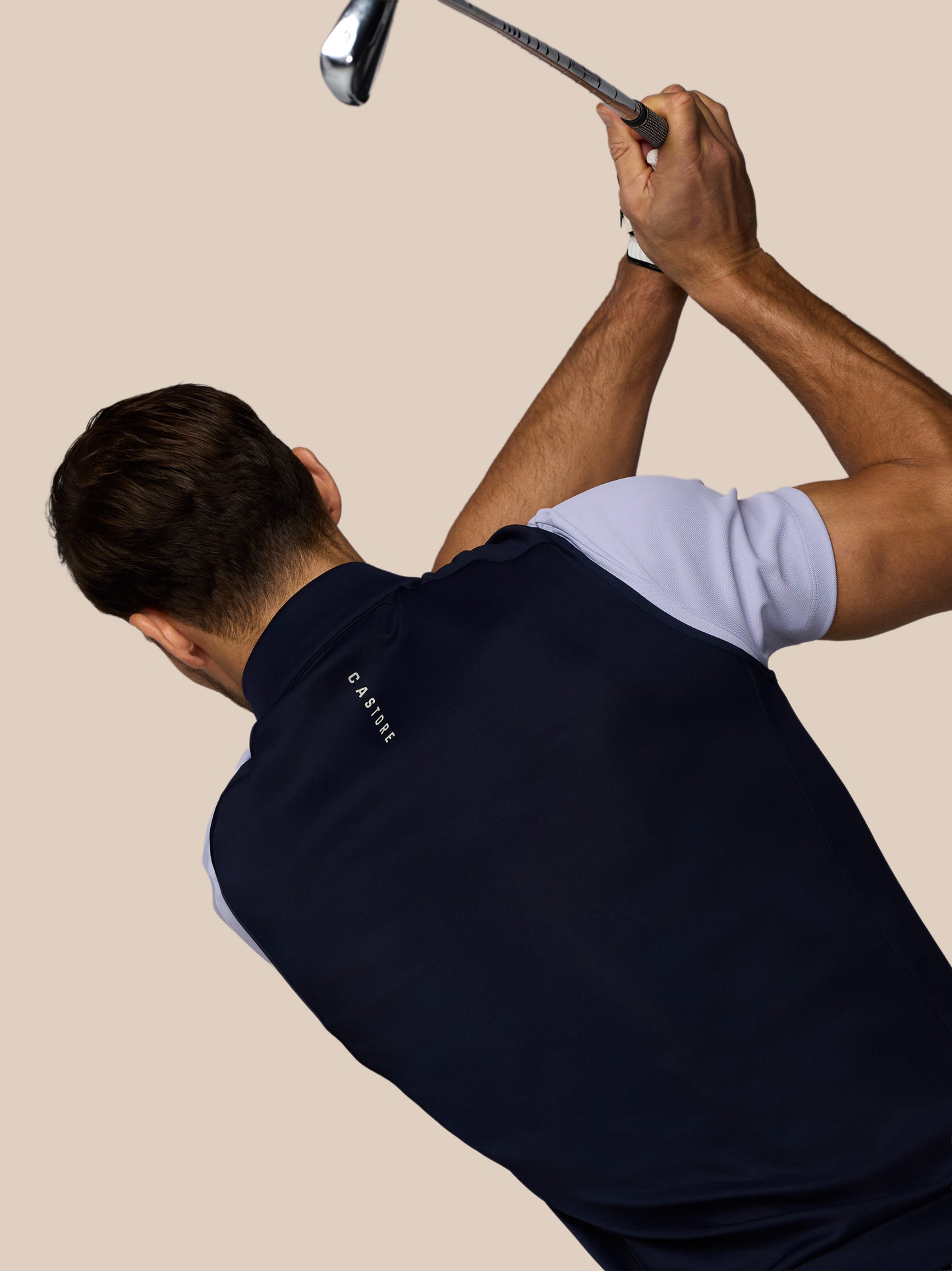 Male golfer wearing a navy Castore golf vest with arms raised in a backswing while gripping a club