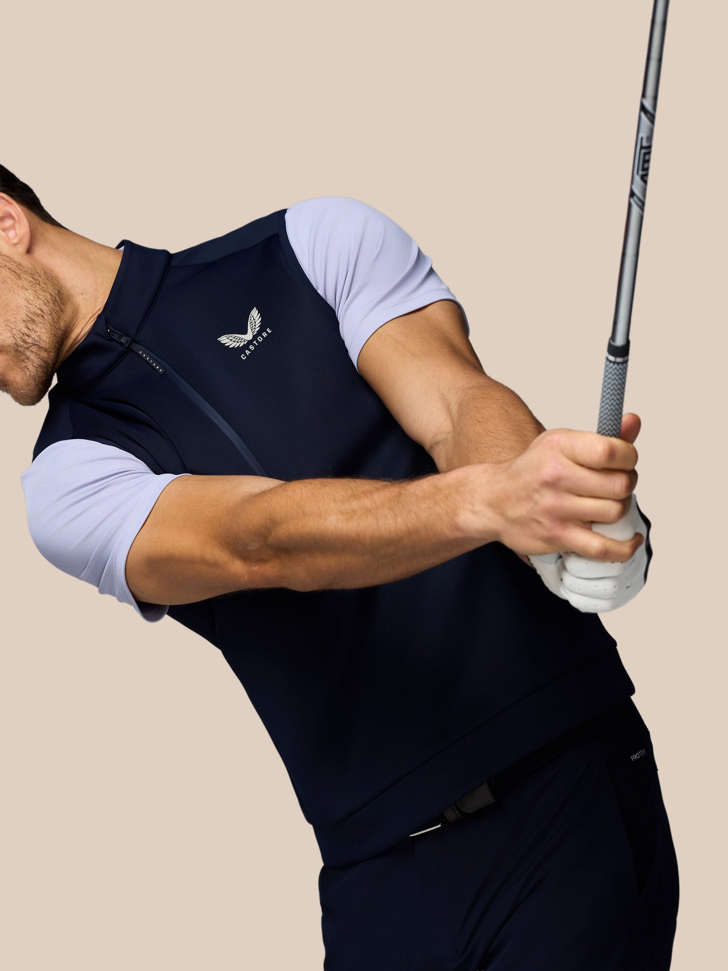 Man wearing navy Castore golf flex half zip gilet with light sleeves, chest logo, gripping a golf club.