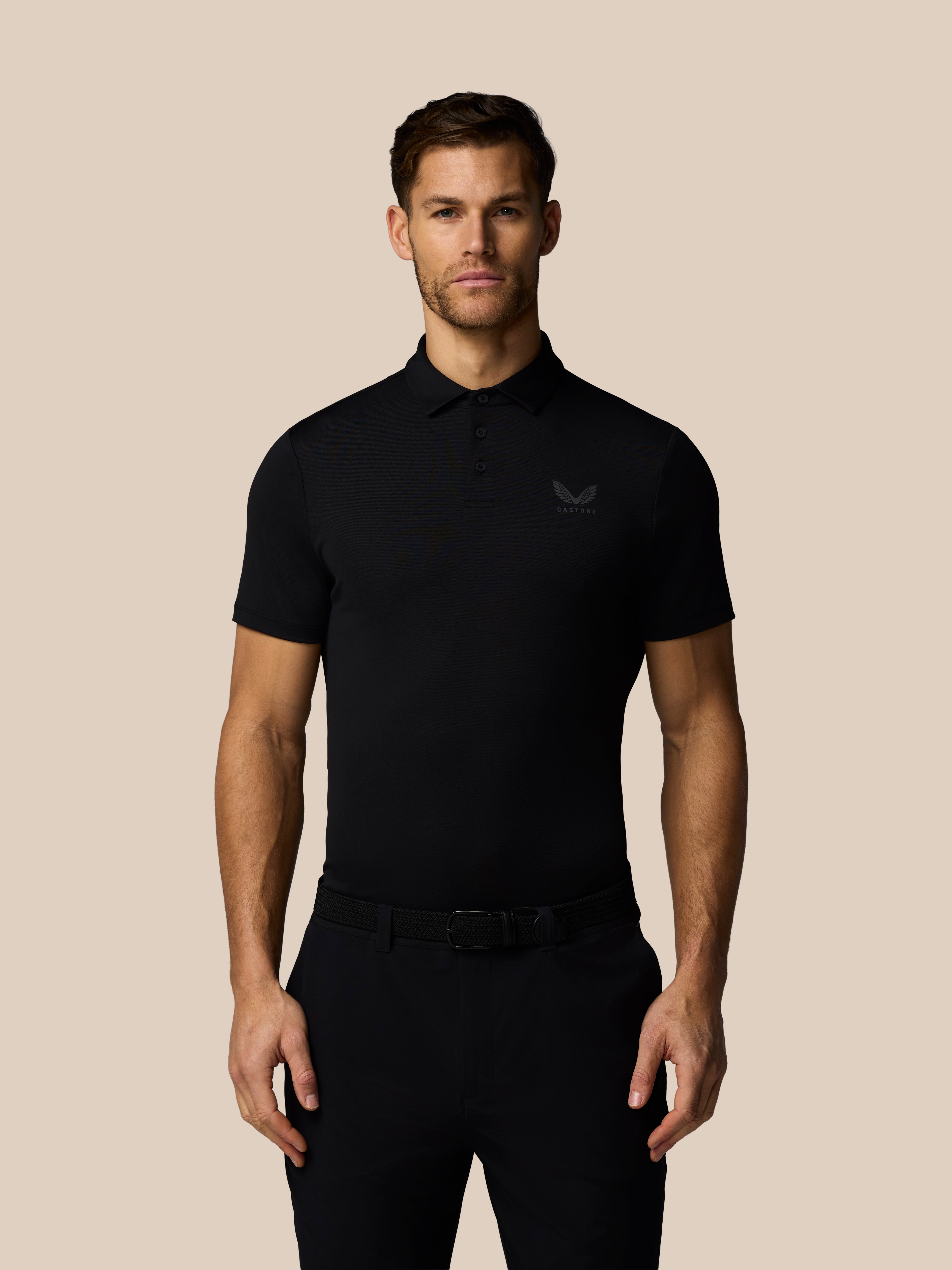 Male model wearing black Men’s Golf Essential polo showing chest Castore logo and moisture-wicking AerTek fabric against beige backdrop