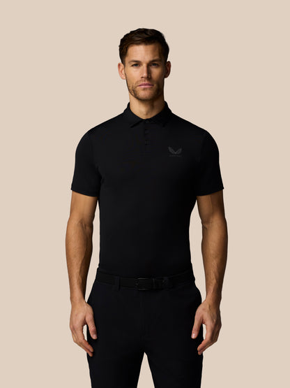 Male model wearing black Men’s Golf Essential polo showing chest Castore logo and moisture-wicking AerTek fabric against beige backdrop