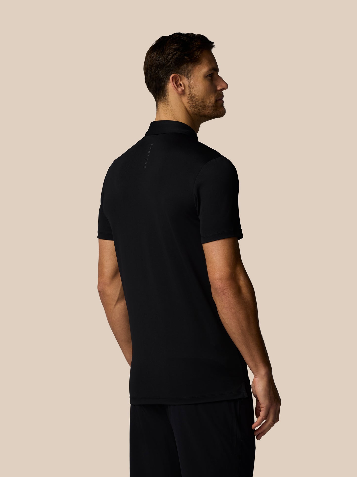 Back view of male model wearing black Castore golf polo with upper back branding against beige backdrop