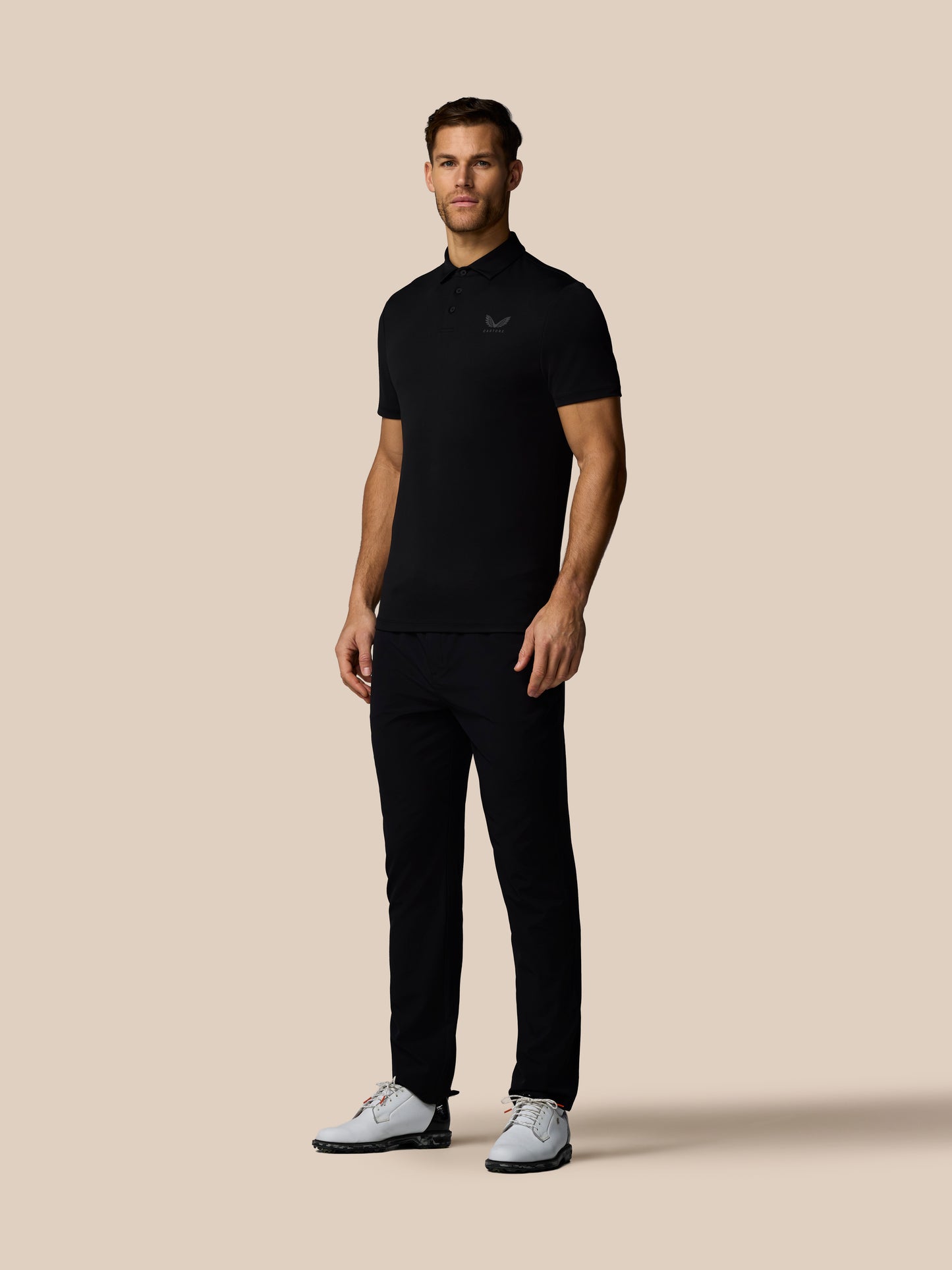 Three quarter rear view of male model in black golf polo with subtle vertical Castore branding along back and white sneakers