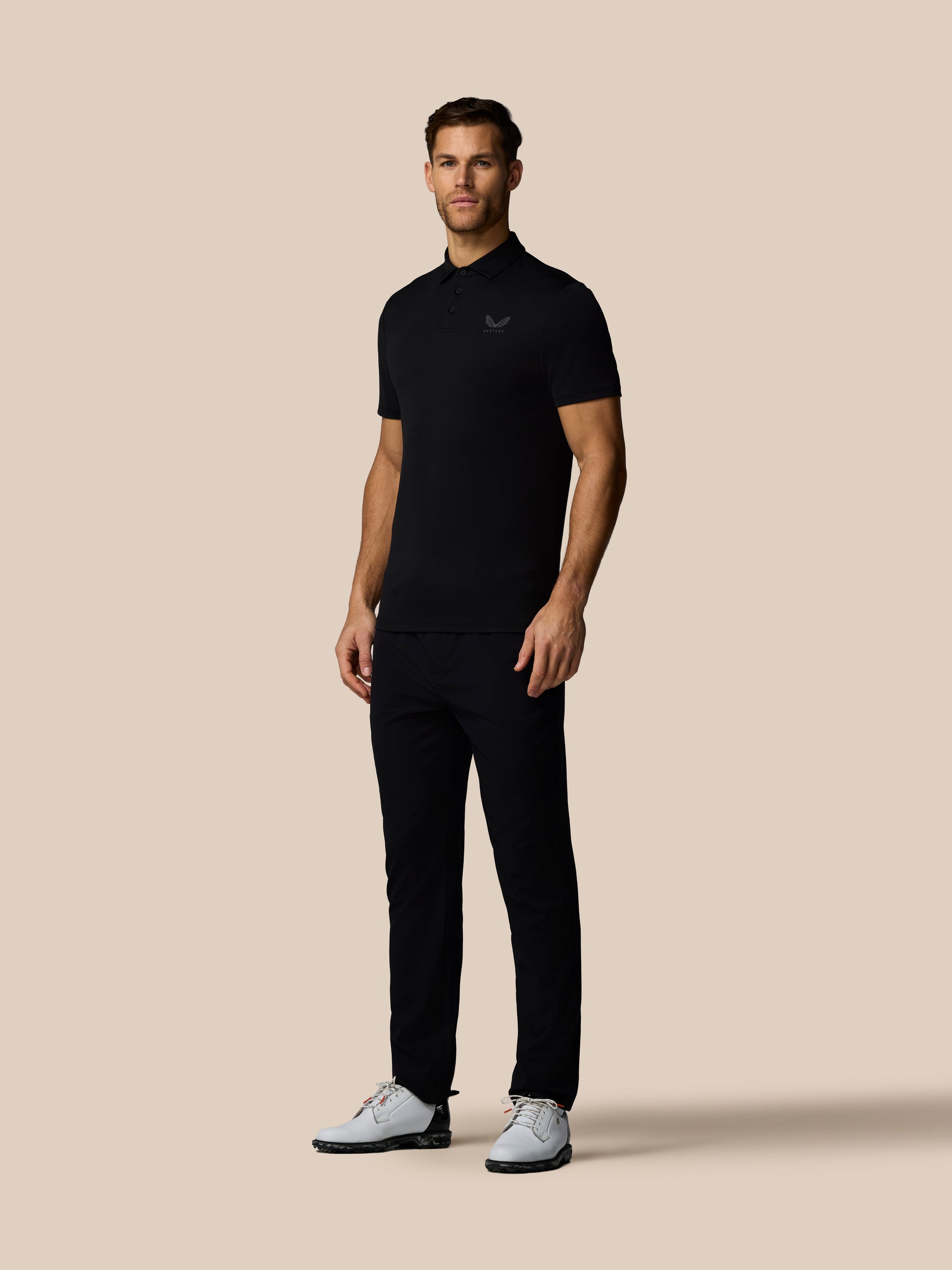 Three quarter rear view of male model in black golf polo with subtle vertical Castore branding along back and white sneakers