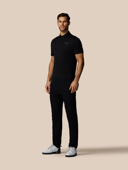 Three quarter rear view of male model in black golf polo with subtle vertical Castore branding along back and white sneakers