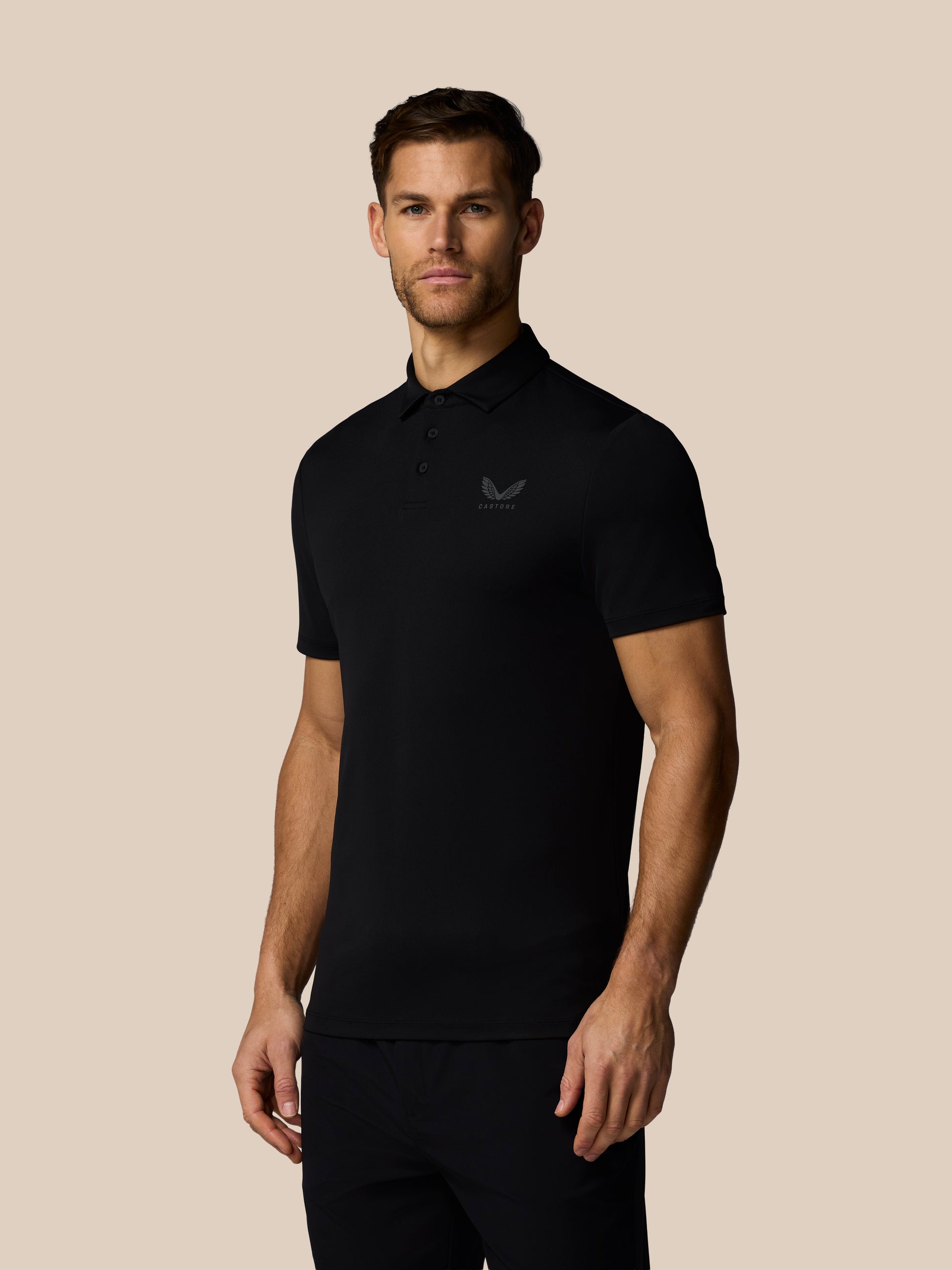 Man wearing black Castore golf polo with left chest logo and back wordmark, standing against a beige background