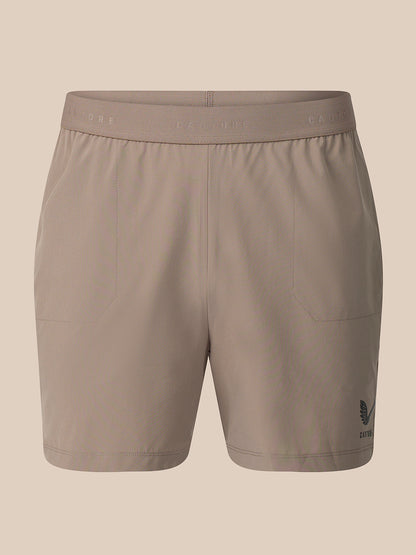 A pair of women's athletic shorts in a solid taupe color, featuring a wide elastic waistband with subtle branding and a small dark logo on the lower left leg, shown against a solid beige background.