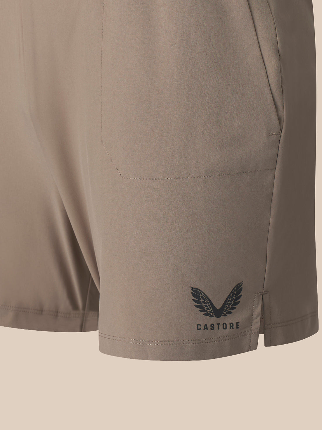 Close-up of a pair of light taupe athletic shorts showing a side pocket and a dark gray Castore wing logo on the lower left leg, set against a solid light beige background.
