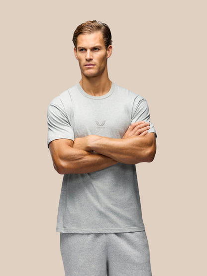 A muscular man wearing a light gray short-sleeve athletic t-shirt and matching gray sweatpants, standing with arms crossed against a solid beige background.