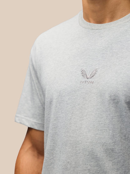 A close-up of a person wearing a light heather gray crewneck t-shirt with a small winged logo and 'CASTORE' text on the left chest, set against a solid beige background.