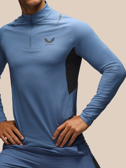A close-up of a man wearing a blue long-sleeve athletic quarter-zip top with black side panels and a gray wing logo on the chest, set against a solid beige background.