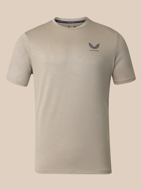 A light beige short-sleeve athletic t-shirt with a subtle ribbed texture, featuring a dark gray wing logo and the brand name 'CASTORE' On the left chest, displayed against a solid beige background.