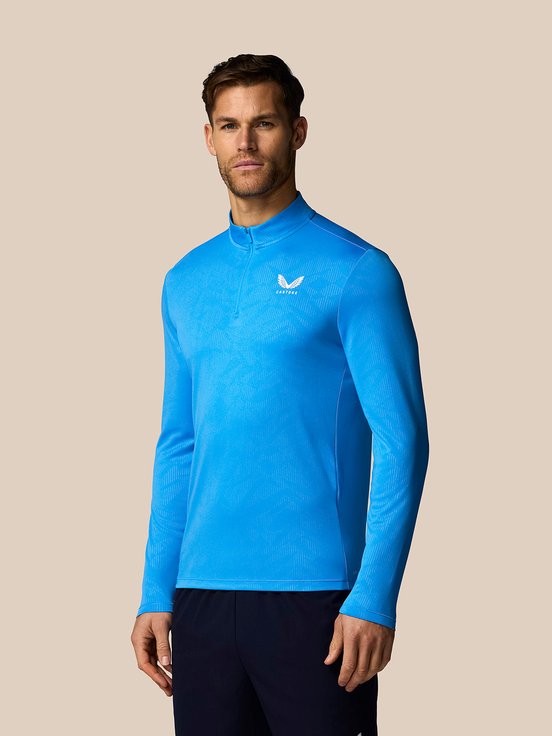 Male model wearing blue quarter zip long sleeve top with subtle all-over print and Castore logo on chest