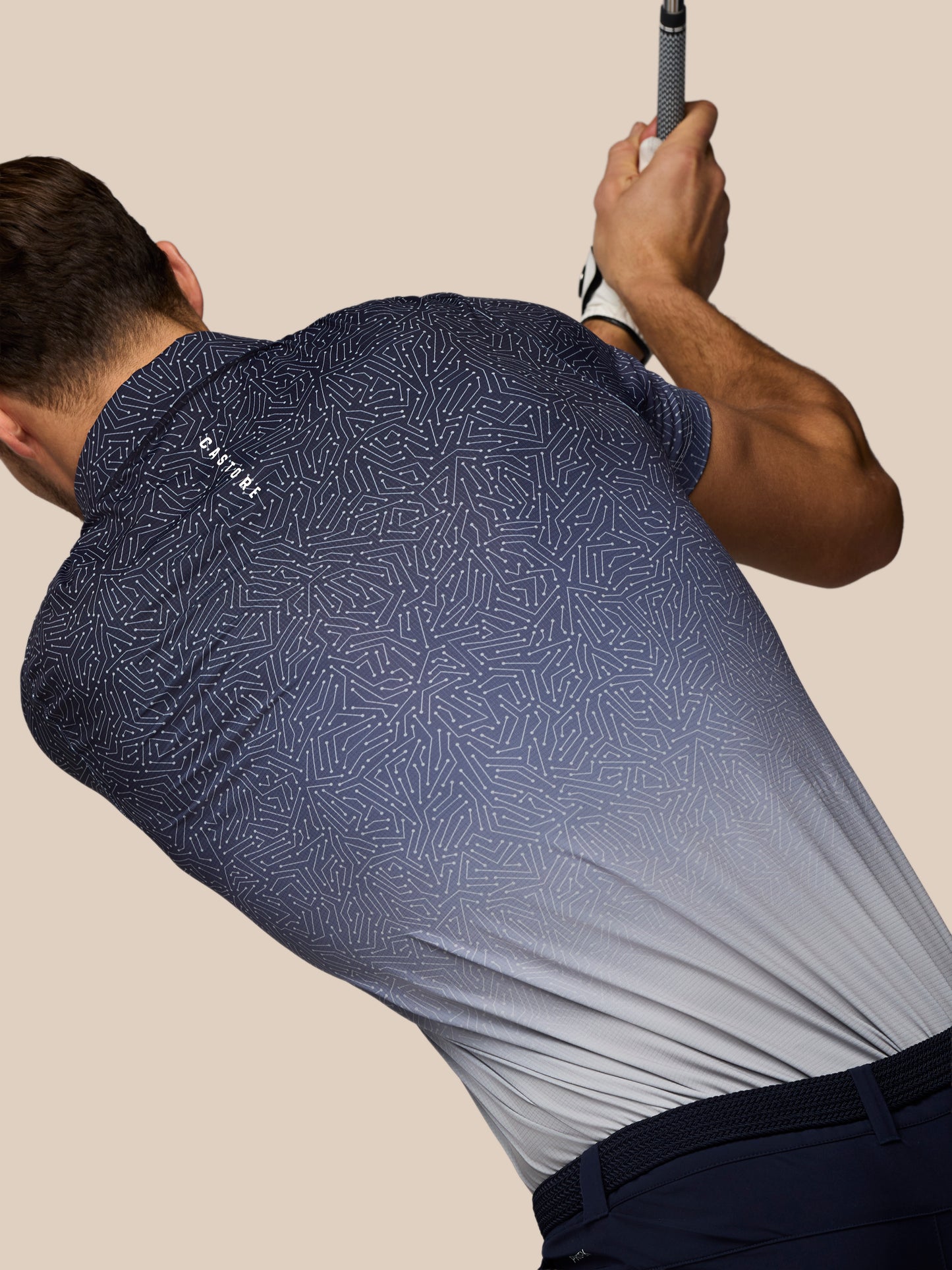 Back view of a golfer wearing a navy map-printed Castore polo during a swing.