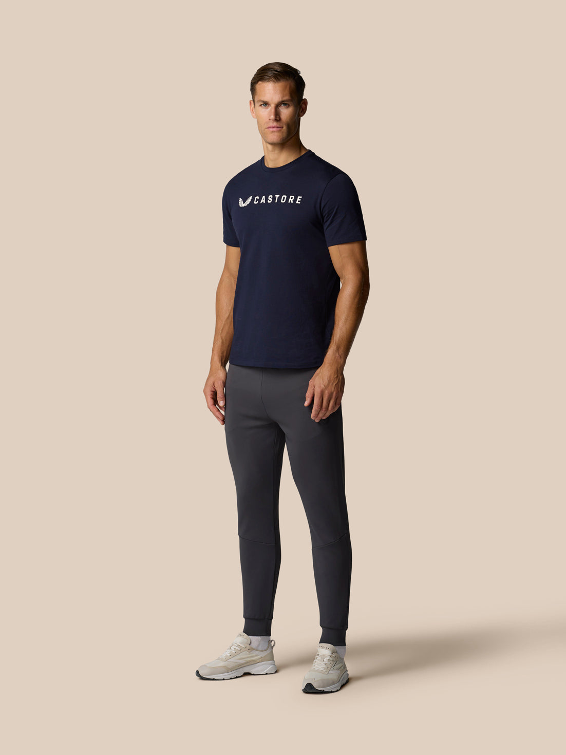 Male model wearing navy Castore graphic lifestyle T shirt with white logo, paired with dark gray joggers.