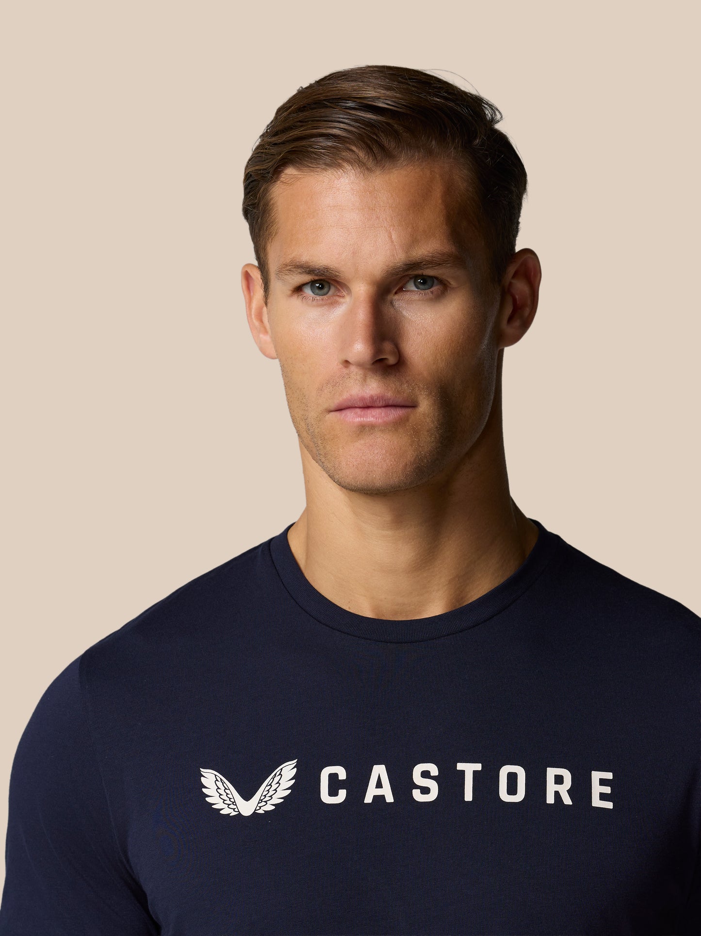 Close-up of a man wearing a navy Castore graphic lifestyle T-shirt with a white wing logo on the chest against a beige background.