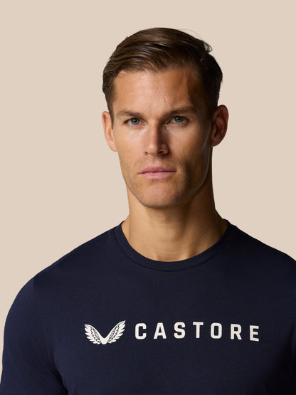 Close-up of a man wearing a navy Castore graphic lifestyle T-shirt with a white wing logo on the chest against a beige background.