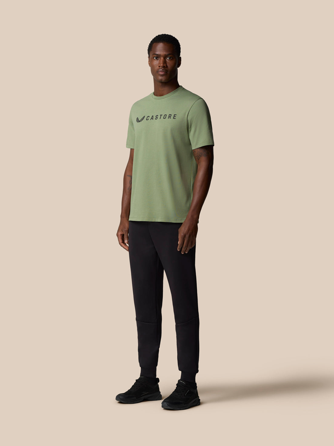 Full body view of a male model wearing thyme green Castore Graphic Lifestyle T Shirt with short sleeves and black pants.