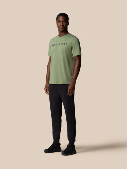 Full body view of a male model wearing thyme green Castore Graphic Lifestyle T Shirt with short sleeves and black pants.