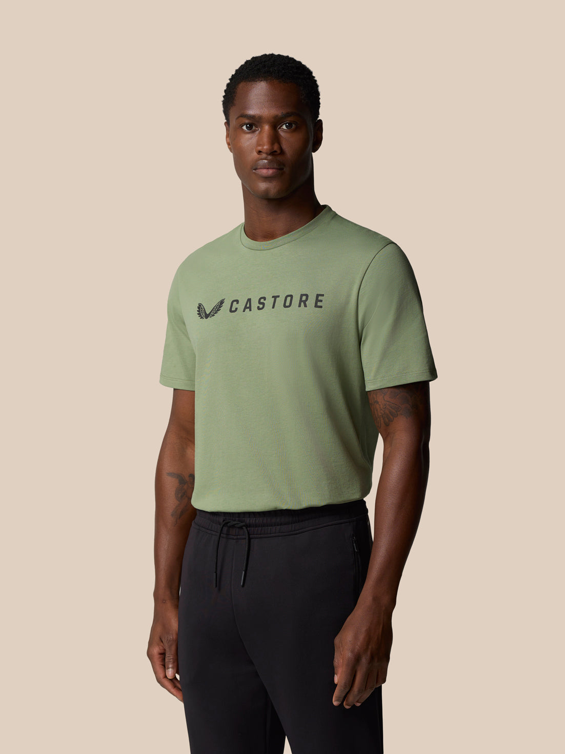 Full length shot of a male model in thyme green Castore Graphic Lifestyle T Shirt with short sleeves and black pants, wordmark on chest.