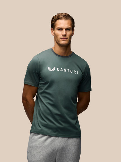 A man wearing a dark green Castore short-sleeve t-shirt with a white logo on the chest, paired with gray sweatpants, set against a solid beige background.