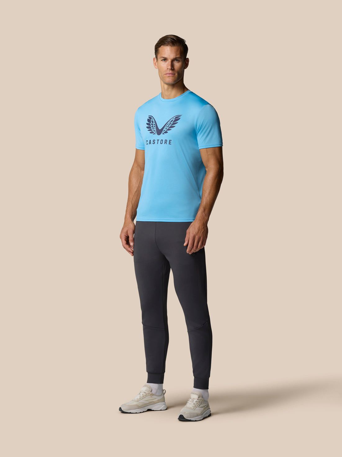 Full-length view of a male model in a light blue Castore Graphic Training T-Shirt with a wing graphic, paired with dark joggers and white sneakers.
