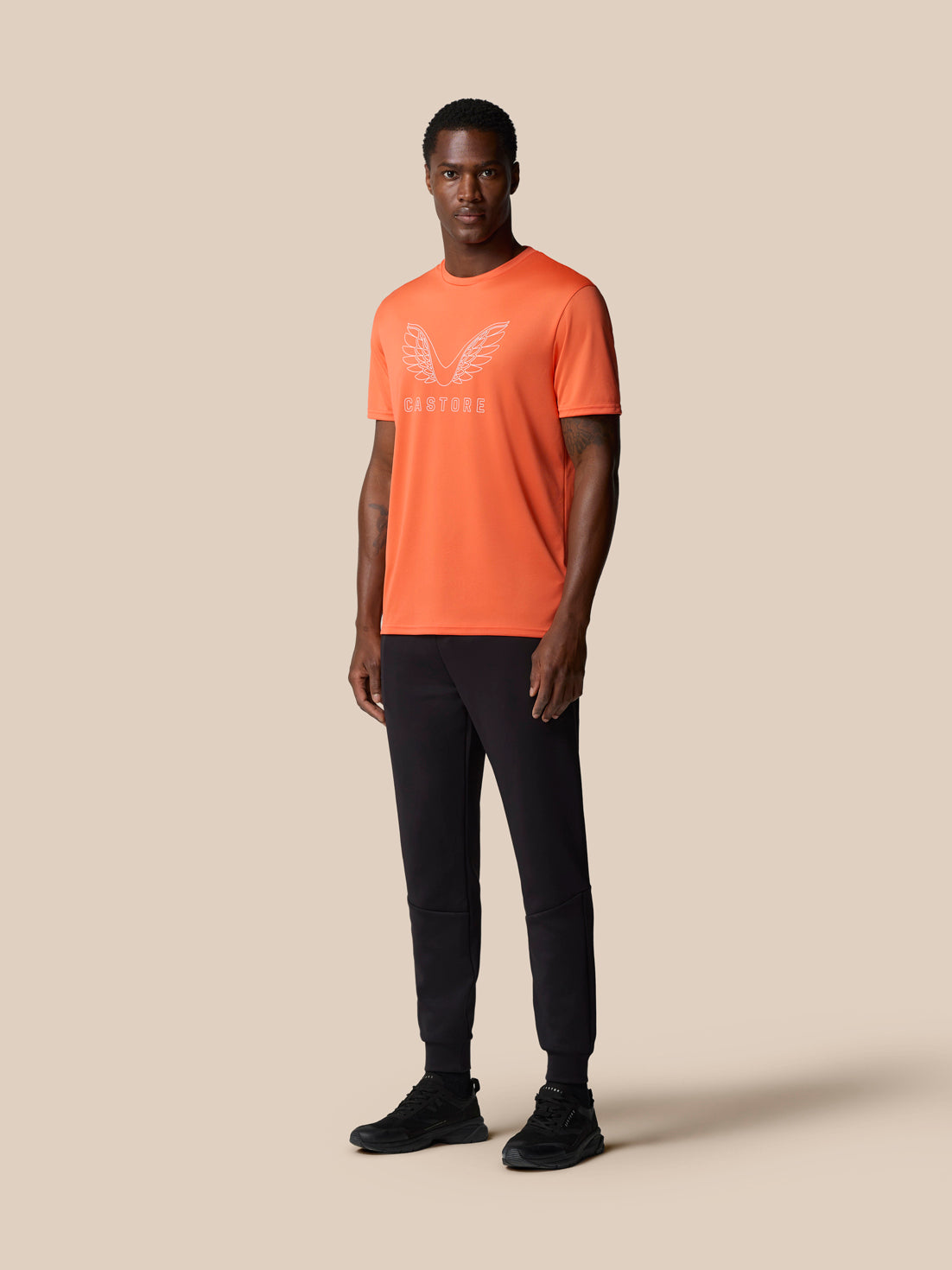 Male model wearing bright orange Castore graphic training T shirt with white wing logo, black joggers and sneakers.