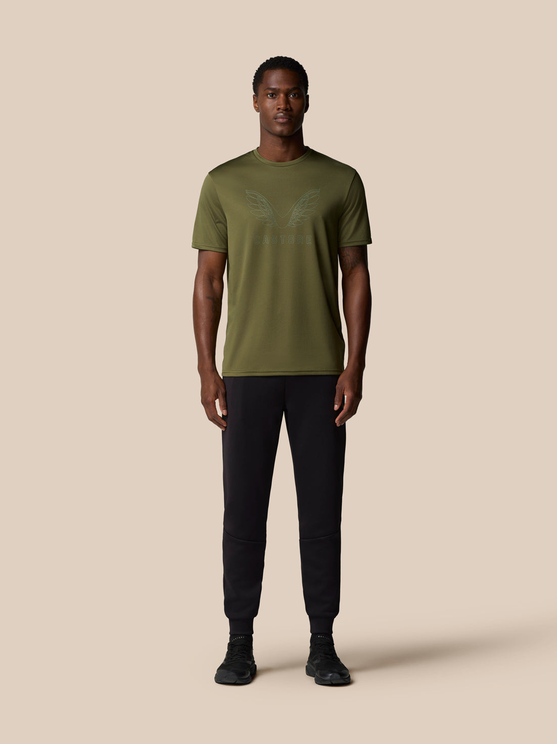 Male model wearing ivy green graphic training T-shirt with wing emblem and Castore logo, paired with black joggers.