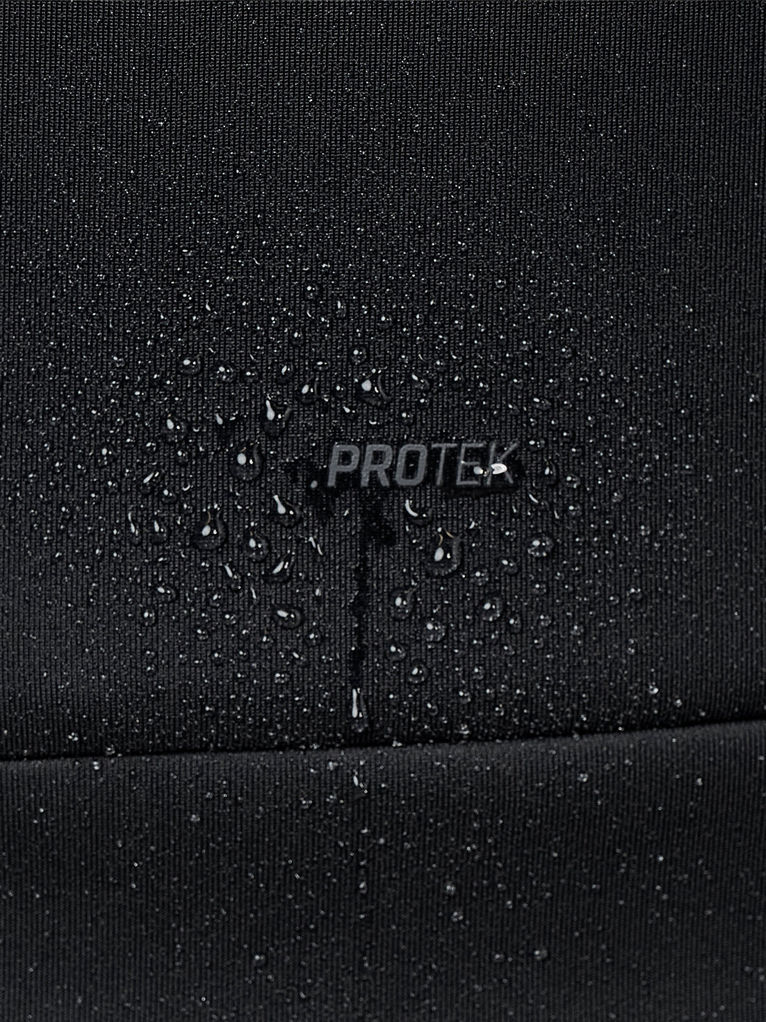 Close-up of a black waterproof fabric with water droplets beaded on the surface and a central 'PROTEK' logo, showing water beading over the text.