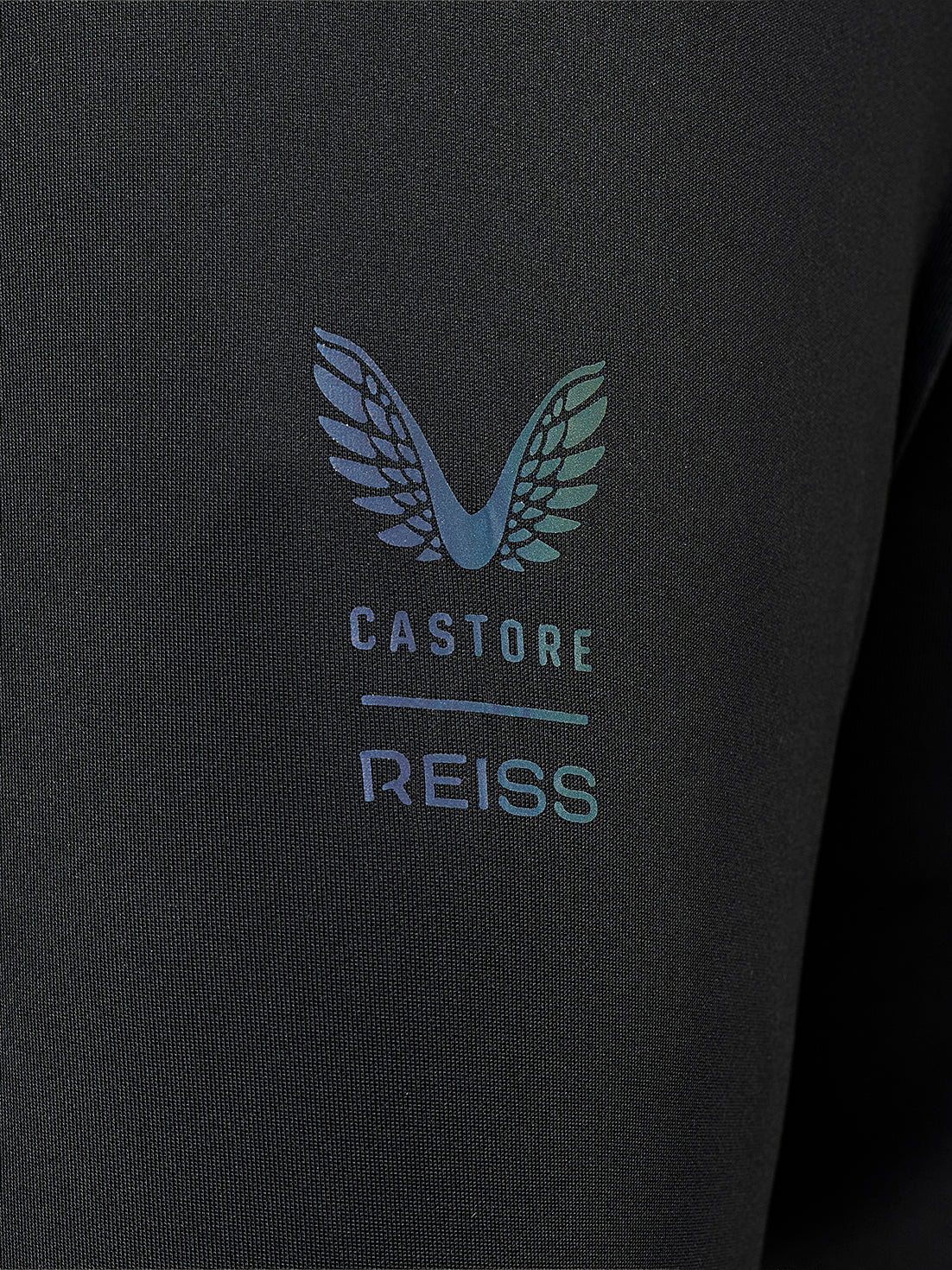 Close-up of a black textured fabric garment featuring a printed winged logo and the text 'CASTORE REISS' in a light blue and teal gradient.