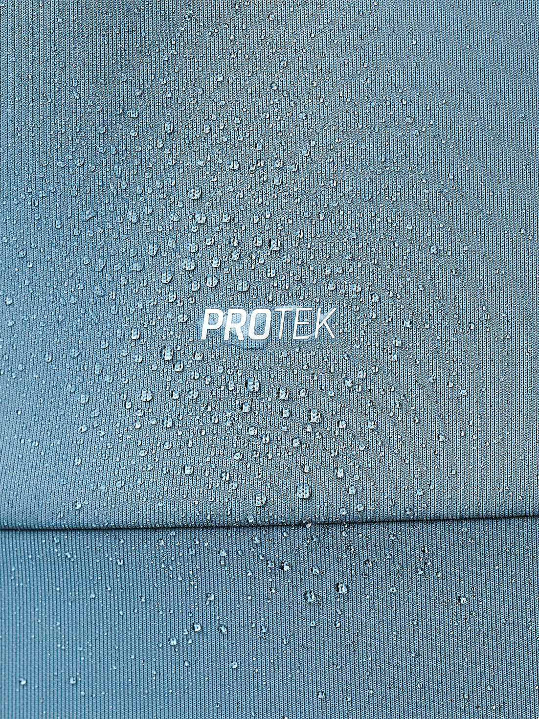 Close-up of a blue-gray textured fabric covered in water droplets, with the word PROTEK printed in white at center. The image demonstrates a water-resistant or protective finish.