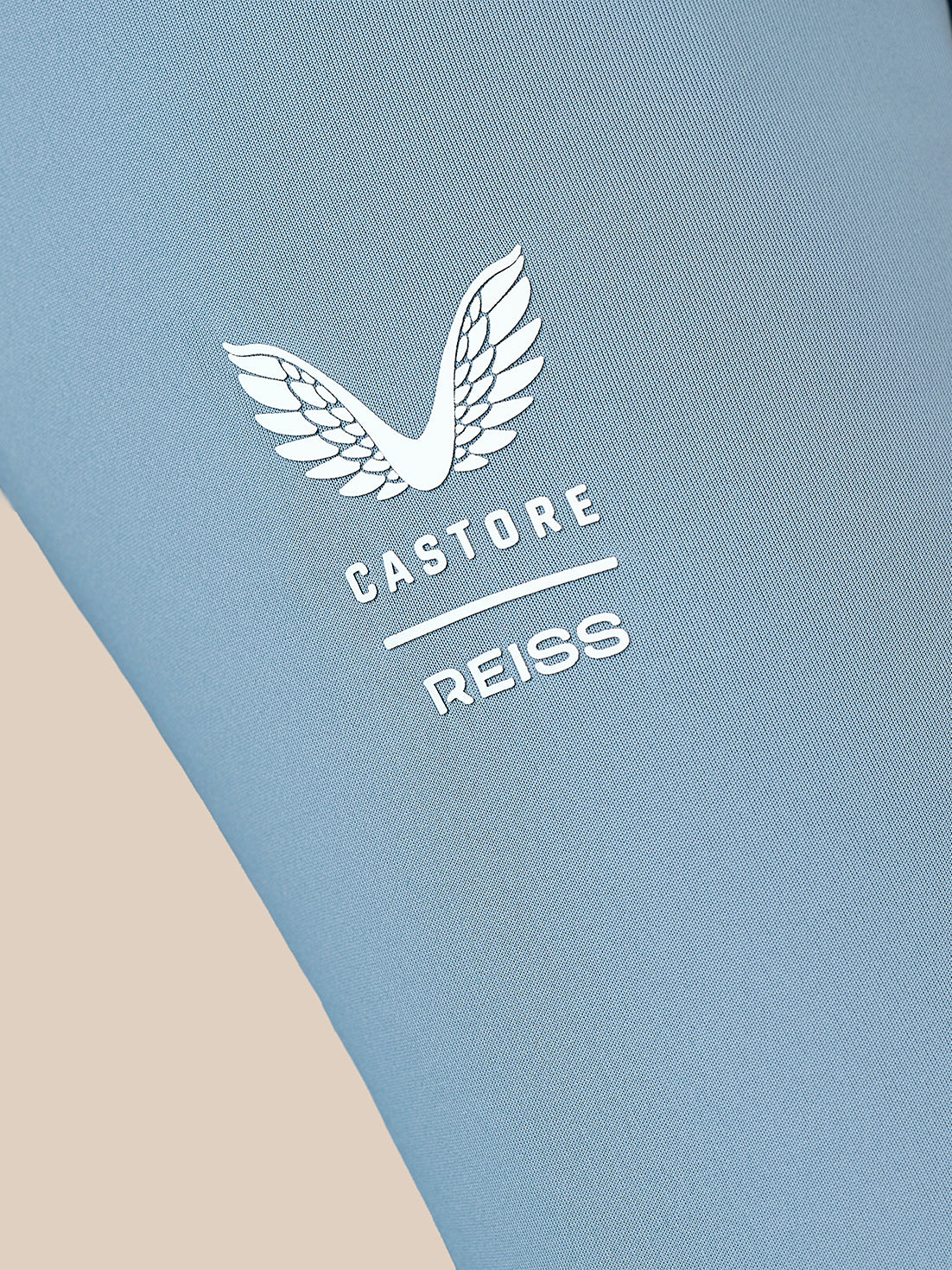 Close-up of a light blue athletic fabric with a white embroidered wing logo and the text 'CASTORE REISS' printed on it.