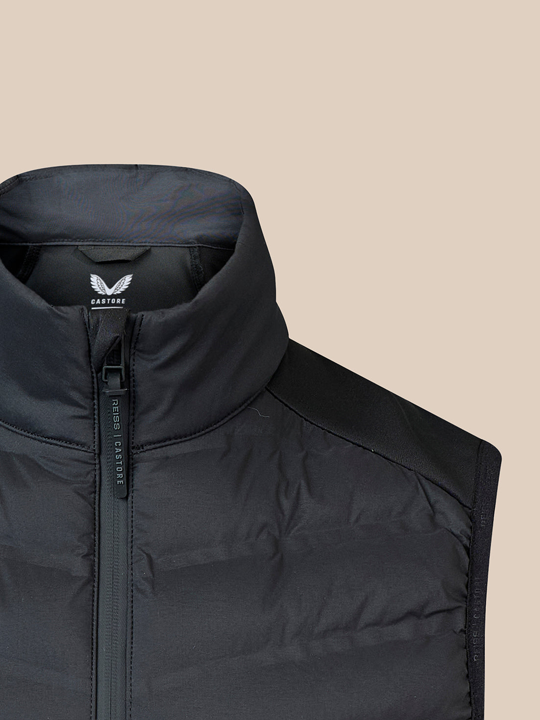 Close-up of a black quilted puffer vest with a stand-up collar and front zipper, shown against a solid beige background. The brand name 'CASTORE' is visible on the inner neck label.