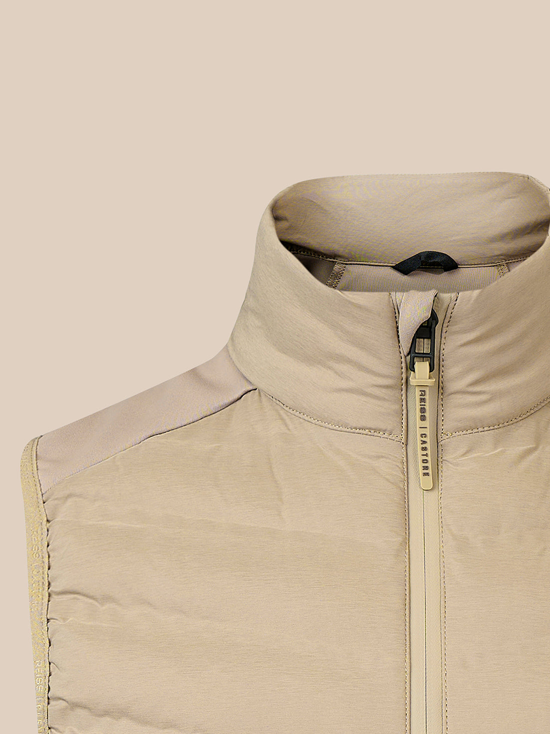 Close-up of a beige puffer vest with a high stand-up collar and a gold-tone zipper pull, shown against a solid beige background.